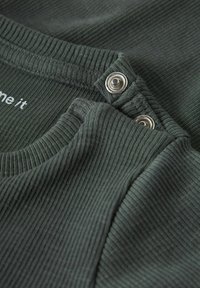 Donkergroene geribbelde stof met een ronde halslijn. Voorzien van zilveren drukknoopsluitingen en subtiele tekstdetails in wit. Zachte textuur.