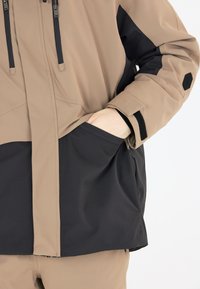 Vattentät jacka i beige och svart tyg, med en rymlig ficka, justerbar huva med dragsnören och kardborreslås vid handledarna.