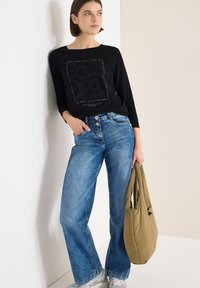 Zwart shirt met lange mouwen en een grafische print, gecombineerd met lichtblauwe jeans met wijde pijpen. Houdt een gewatteerde olijfgroene tas vast. Casuele schoenen zichtbaar.