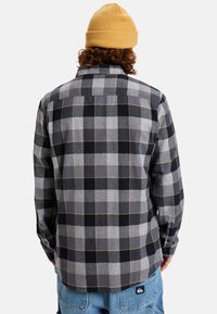 Gray plaid shirt with black and white check patterns, long sleeves, button-down collar, and a yellow stitched accent. Worn with a yellow beanie.