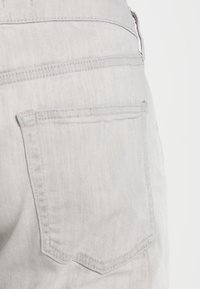 Vaaleanharmaat farkut, joissa on takatasku kaksinkertaisella ompeleella ja hienovaraisella tekstuurilla. Kangas vaikuttaa pehmeältä ja siististi viimeistellyltä.