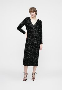 Sieviete, kas ģērbusies melnā garajā piedurkņu sequin midi kleitā un melnās striķu augstpapēžu sandalēs, stāv uz vienkārša fona.