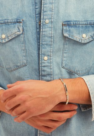 Sølv- og guldfarvet armbånd med et sammenkædet design og sorte accenter, båret på håndleddet over en lyseblå denimskjorte.