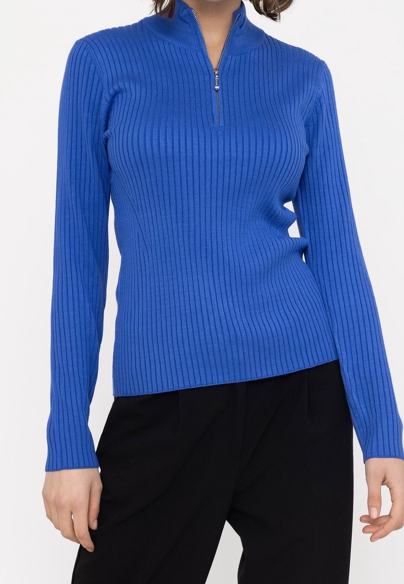 Ribbad blå långärmad topp med dragkedja i kragen. Materialet verkar slätt och stretchigt, åtsittande genom torselet, med en ren finish.