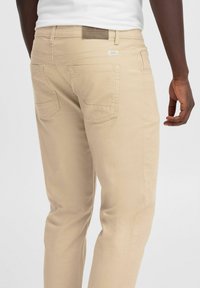 Pantaloni in cotone beige con taglio dritto, caratterizzati da una consistenza liscia, tasche posteriori e un discreto etichetta logo sulla vita.