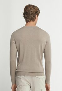 Beige stickad tröja med lång ärm, rund halsringning och ribbade muddar med en mjuk textur. Vy bakifrån.