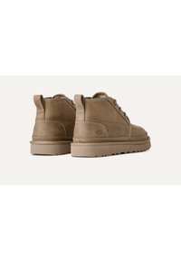 Botas de tobillo de ante marrón con suela de goma plana, punta redonda y un diseño mínimo de cordones; cuenta con la marca UGG en el lateral.