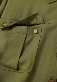 Olivegrüner Stoff mit strukturierter Oberfläche verfügt über eine aufgesetzte Tasche und zwei Messingdruckknöpfe, die funktionale Designelemente hervorheben.