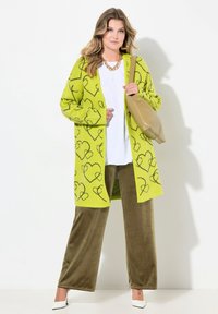 Cardigan a maglia di colore verde neon con motivi a cuore neri, abbinato a pantaloni in velluto verde oliva e scarpe a tacco bianche. Tiene una borsa beige.