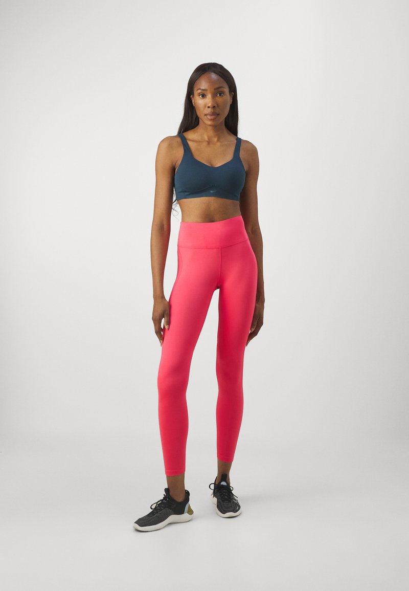 Mujer de pie vistiendo un sujetador deportivo azul marino, leggins rosas brillantes y zapatillas deportivas negras sobre un fondo blanco liso.