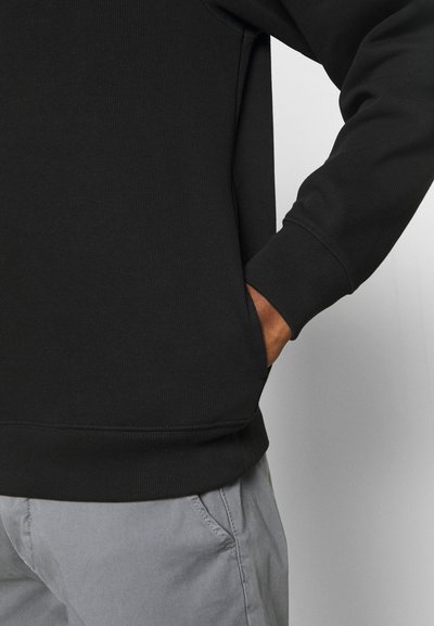 Sweatshirt noir à côtes avec une coupe décontractée, présentant des manches longues et une poche sur le côté. Associé à un pantalon gris clair.