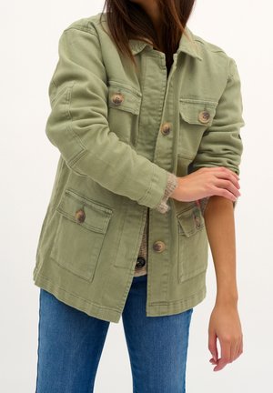 Personne ajustant la manche retroussée d'une veste boutonnée vert olive portée sur un pull beige et un jean bleu, sur un fond uni.