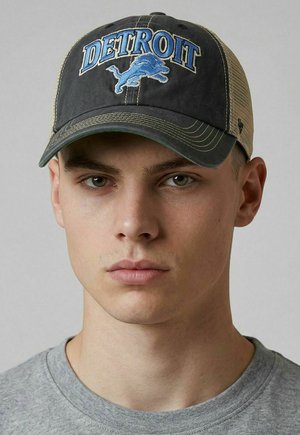 Young man wearing a black and beige Detroit Lions baseball cap and a gray crew neck shirt against a plain background.