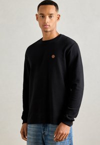 Mand iført en sort langærmet skjorte med et lille brunt rundt logo på brystet, parret med blå jeans, stående mod en ensfarvet baggrund.