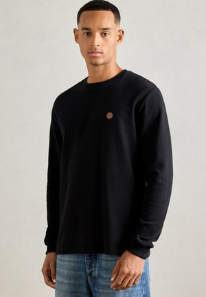 Man wearing a black long-sleeve shirt with a small brown circular logo on the chest, paired with blue jeans, standing against a plain background.