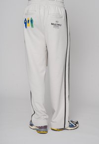 Weiße Jogginghose mit schwarzen Seitenstreifen und gestickten Figuren in Blau, Gelb und Grün auf der linken Tasche; mit einem Kordelzugbund.