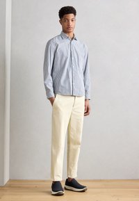 Stripet skjorte med knapp, lyseblå og hvit; kremfargede bukser; svarte slip-on sko; tregulv, minimalistisk bakgrunn.