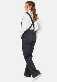 Dunkelblaue Denim-Latzhose mit weit auslaufenden Beinen, verstellbaren Trägern und Gesäßtaschen. Getragen über einem weißen Langarmshirt und weißen Turnschuhen.
