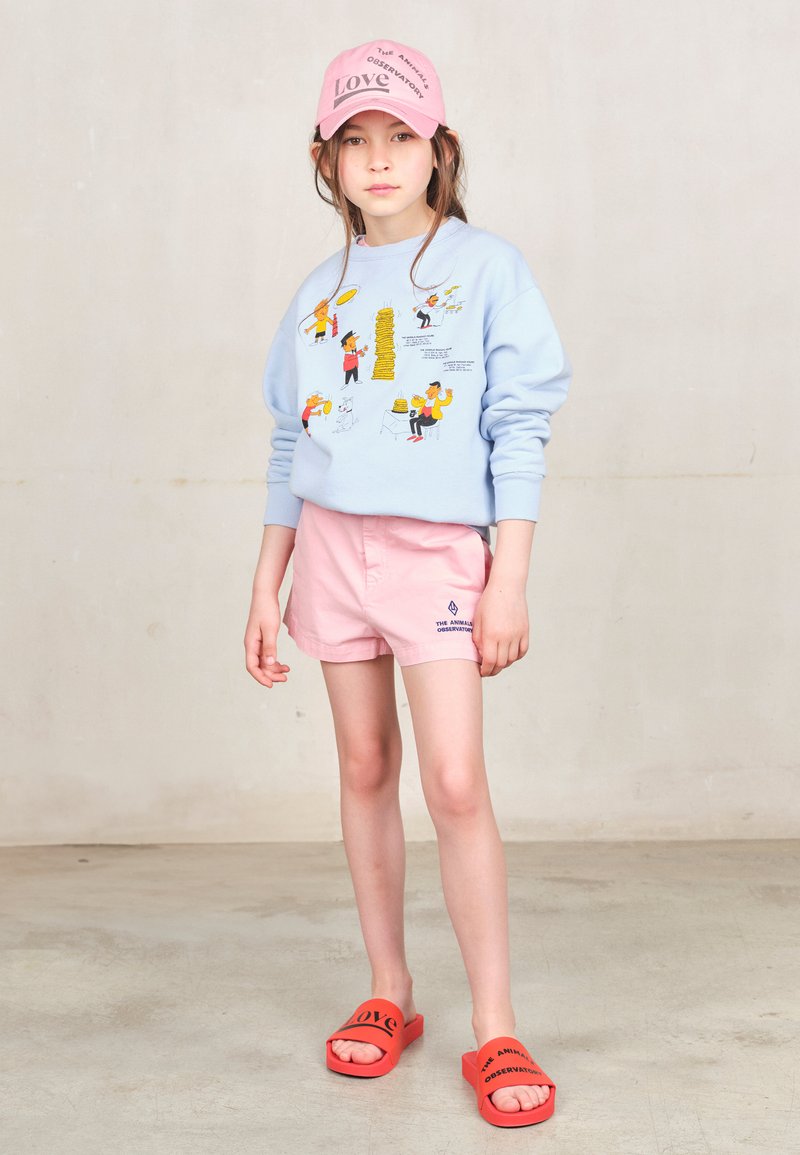 Felpa azzurra chiara con stampe di cartoni animati colorate, pantaloni corti rosa e ciabatte rosse con testo. Materiali morbidi, vestibilità casual e design semplice.