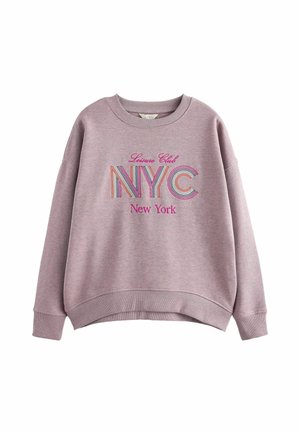 Femme portant un sweat-shirt NYC couleur lavande, un pantalon imprimé léopard et des baskets bordeaux, debout avec une main dans la poche sur un fond beige.