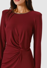 Vestito bordeaux a maniche lunghe con un dettaglio a nodo intrecciato in vita e un accento metallico lungo la scollatura, realizzato in tessuto liscio.