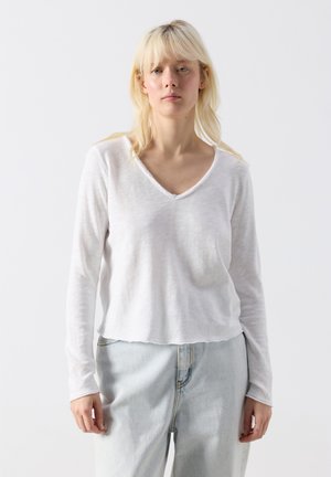 Pikajuukseline naine, kes kannab valget pika varrukaga V-kaelusega topi ja helesiniseid teksapükse, seistes ühevärvilise valge tausta ees.