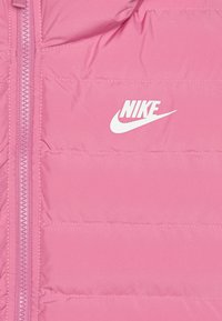 Roze puffer jakna s vertikalnim prešanim dizajnom, koja ima prednji zatvarač i bijeli Nike logo na lijevoj strani prsnog koša.