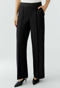 Black wide-leg trousers with a smooth texture, featuring a tonal elastic waistband and front pleats, worn with pointed black heels.