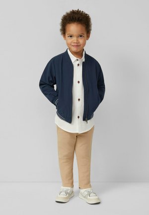 Junges Kind steht mit Händen in den Taschen, trägt eine marineblaue Jacke, weißes Hemd mit großen Knöpfen, beige Hose und weiße Turnschuhe vor einfachem Hintergrund.