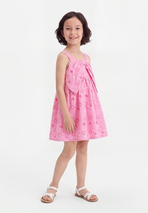 Κορίτσι που στέκεται και χαμογελά φορώντας αμάνικο ροζ φόρεμα με λουλουδένια σχέδια και λευκά σανδάλια σε λευκό φόντο.