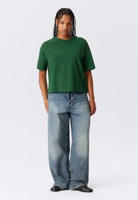 T-shirt verde a maniche corte con vestibilità rilassata, abbinato a jeans blu chiaro a gamba larga. Stivaletti neri completano l'outfit.