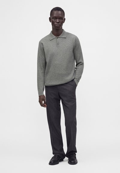 Homme portant un polo en maille gris à manches longues, un pantalon gris foncé et des chaussures de ville noires sur un fond blanc uni.