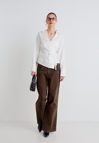 Λευκό πουκάμισο τύπου wrap με μακριά μανίκια, συνδυασμένο με καφέ παντελόνι φαρδύ στη μπότα. Αξεσουάρ περιλαμβάνουν μια μαύρη τσάντα με τρουκς και μυτερές μαύρες γόβες.