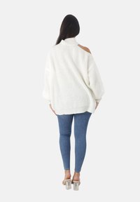 Femme aux longs cheveux noirs portant un pull en tricot blanc oversize avec une épaule dénudée, un jean skinny bleu et des sandales à talons transparents, de dos.