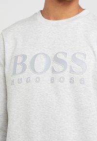 Hellgrauer Rundhalsausschnitt-Pullover mit erhabenem "BOSS HUGO BOSS"-Logo auf der Brust, getragen von einer Person, die vom Hals bis zum Oberkorb zu sehen ist.