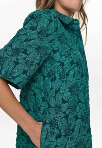 Cămașă verde cu un model floral, cu guler, mâneci scurte și un material texturat. Are o pungă laterală pe partea stângă.