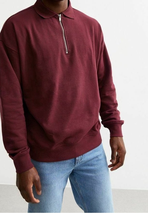 Bordeaux pullover sweatshirt met een ritssluiting kraag, lange mouwen en ribgebreide manchetten. Gecombineerd met lichtblauwe jeans, weergegeven tegen een lichte achtergrond.