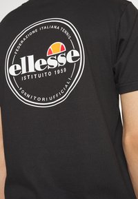 T-shirt noire avec un logo circulaire portant "ellesse" en blanc, accentué par des détails rouges et jaunes, et le texte "Fornitori Ufficiali" en dessous.