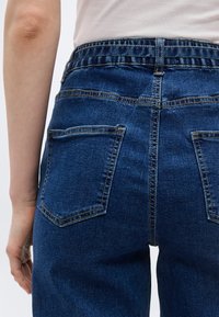 Πίσω όψη ατόμου που φοράει ψηλόμεσο σκούρο μπλε τζιν με ελαστική μέση και εμφανείς πίσω τσέπες, με το χέρι χαλαρό στο πλάι.