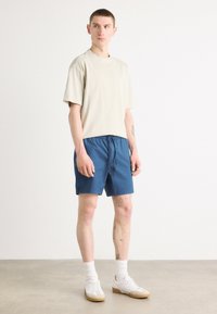 T-shirt beige oversize, short bleu à taille élastique, chaussettes blanches et baskets blanches avec semelle beige. Texture en coton lisse, coupe décontractée.
