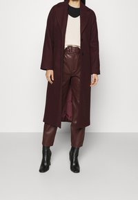 Femme portant un long manteau bordeaux, un pantalon en cuir ceinturé, un pull en tricot crème, des bottes noires, debout devant un fond blanc uni.