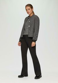 Grijze textuurjacket met zilveren knopen, zwarte gebreide top met een geruit patroon en zwarte flared broeken, afgemaakt met glanzende zwarte schoenen.