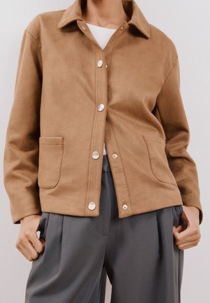 Giacca marrone in materiale scamosciato con colletto, bottoni a pressione argentati e due tasche frontali, abbinata a pantaloni grigi.