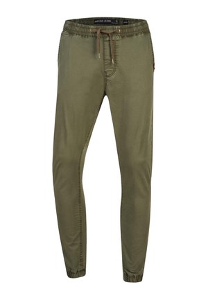 Homme portant un pantalon de jogging vert olive avec taille à cordon et poignets élastiques, associé à des baskets blanches, debout avec une main dans la poche.