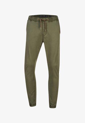 Homme portant un pantalon de jogging vert olive avec taille à cordon et poignets élastiques, associé à des baskets blanches, debout avec une main dans la poche.