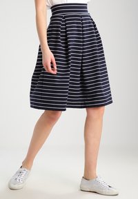 Gonna a righe blu navy con strisce orizzontali bianche, design plissettato e vita alta. Indossata con sneaker bianche, che mostrano un look casual.