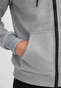 Sudadera gris con cremallera, puños acanalados y un bolsillo frontal. La tela parece suave y texturizada, con una cremallera negra.