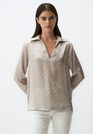 Blusa di un leggero taupe con pois bianchi, scollatura a V e maniche lunghe. Realizzata in un tessuto trasparente e morbido, presenta un design classico del colletto.