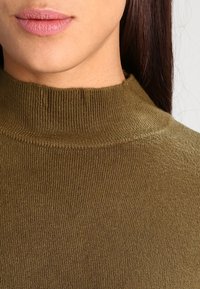 Maglione verde oliva a coste con collo alto. Tessuto morbido e design aderente, caratterizzato da un collo alto e una silhouette avvolgente. Senza fantasie né accessori.
