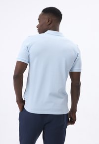 Helles blaues Poloshirt mit klassischem Kragen und kurzen Ärmeln. Der Stoff wirkt glatt mit einer figurbetonten Passform, kombiniert mit dunkelblauen Hosen.